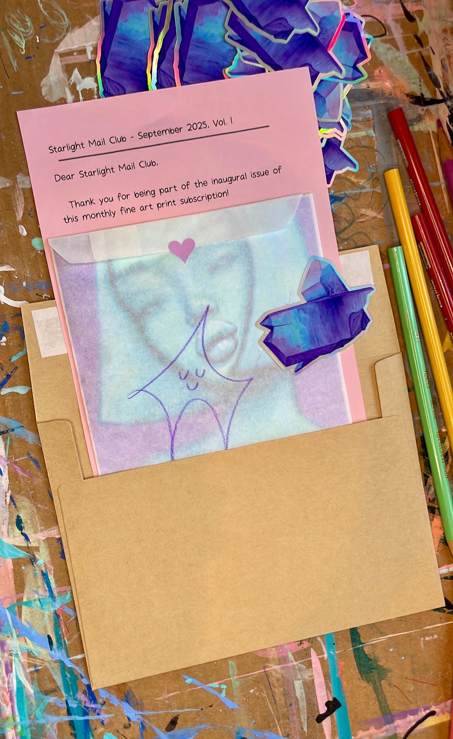 Artistic card with a pink envelope and colorful stationery items on a textured surface